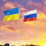 Ukrainian and Russian flags against colorful sunset sky.