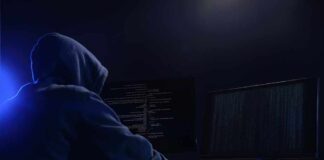 Hooded figure using dual monitors in dark room.
