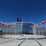 Flags outside NATO headquarters building under clear blue sky.