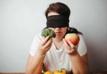 Blindfolded person holding broccoli and an apple.
