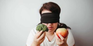 Blindfolded person holding broccoli and an apple.
