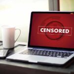 Laptop screen displaying CENSORED beside coffee and phone
