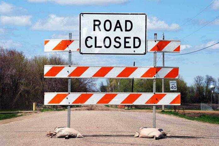 A Road Closed sign with orange and white barriers on a rural road