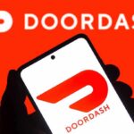 A hand holding a smartphone displaying the Doordash logo against a bright red background