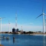 Wind turbines and a communication tower near a body of water under a clear blue sky
