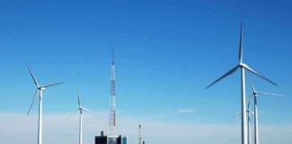 Wind turbines and a communication tower near a body of water under a clear blue sky