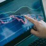 Hand pointing at stock market graph on laptop screen.
