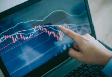 Hand pointing at stock market graph on laptop screen.