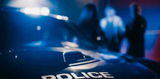 Drive-Thru SHOOTING Mayhem: Bullet Lodged in Dad’s Head Police car with blurred figures in the background.