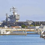 US Shipyards in Peril: TRUMP’S NIGHTMARE REVEALED A large naval aircraft carrier docked in a harbor with smaller boats in the foreground