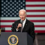 White House Blacklist Targets Leftist Voices Man speaking at podium with US flag background.