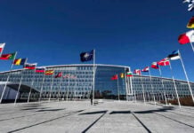 Peace Deal Dangles $300B — What’s The Catch? Flags outside NATO headquarters building under clear blue sky.