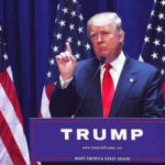 America Isn’t Optional for Newcomers Man speaking at podium with USA flags behind him.