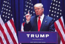 America Isn’t Optional for Newcomers Man speaking at podium with USA flags behind him.