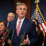 Long Hospital Stay Leaves Voters Waiting Man speaking at a press conference with American flags.