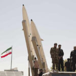 Iran’s Power Transition Faces New Mystery Military personnel beside missiles and Iranian flag.