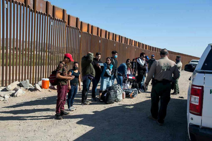 Group of people standing by a border fence with officers and vehicles