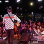 Guitarist performing on stage facing a cheering crowd