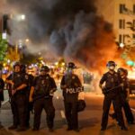 Suspect in Custody After Fiery Attack Outside NYC Christian Jones and Teddy Williams among police line at night protest with fire and smoke