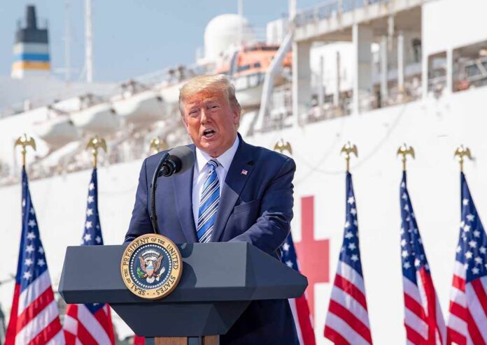 man in suit speaking at a podium with flags and a ship behind