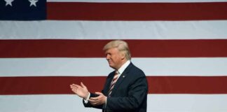 Donald Trump clapping on stage before a large American flag