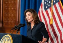 Kathy Hochul speaking at a podium with microphones and a U.S. flag behind her