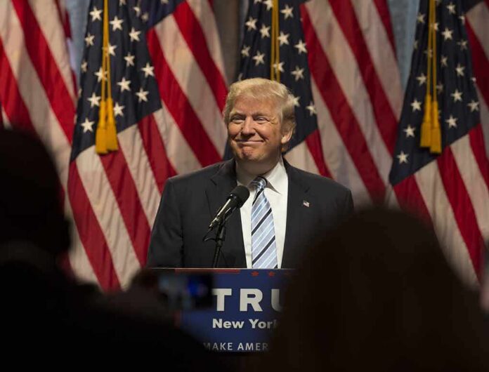 shutterstock_453281794 Man in suit speaking at podium with American flags behind