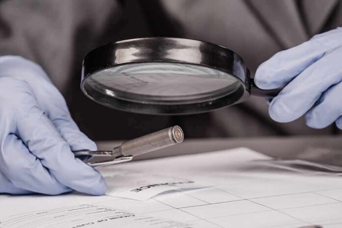Gloved hands examine bullet casing with magnifying glass